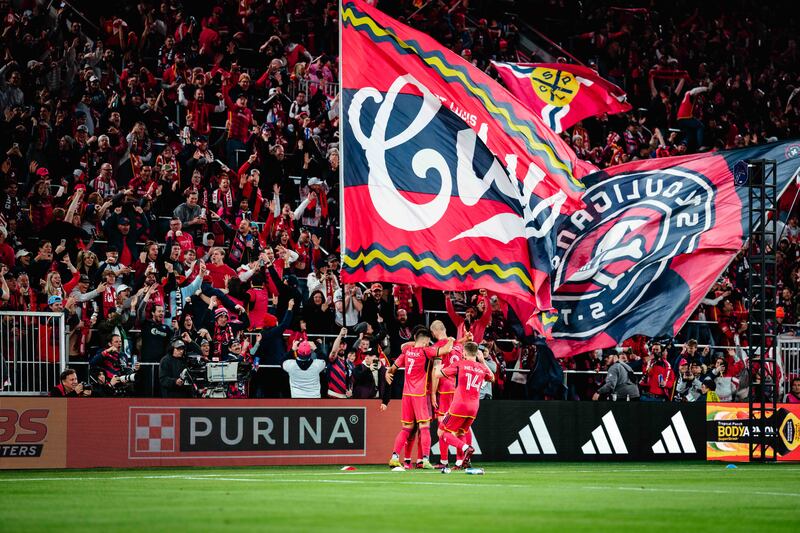 Tomás Ostrák, João Klauss and John Nelson stand hugging each other in front of the CITY SC crowd who are waving CITY flags.